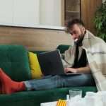 a man on a laptop, with a thermometer in his mouth, wearing red socks and sitting on a couch