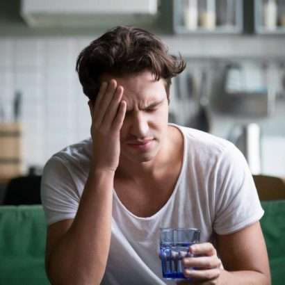 a man with a typical hangover, drinking water and holding his head in his kitchen