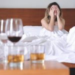 a woman with various alcoholic drinks strewn about her nightstand, she is still in bed and holding her head