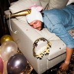 a man wearing a unicorn hat, still passed out on a couch surrounded by balloons, a crown and a beer clutched in his hand