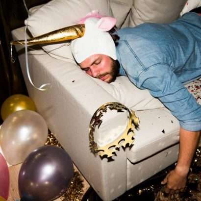 a man wearing a unicorn hat, still passed out on a couch surrounded by balloons, a crown and a beer clutched in his hand